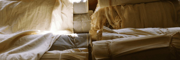Linen fabric stacks in natural cream, beige, and striped in soft sunlight, evoking timeless luxury textiles Header