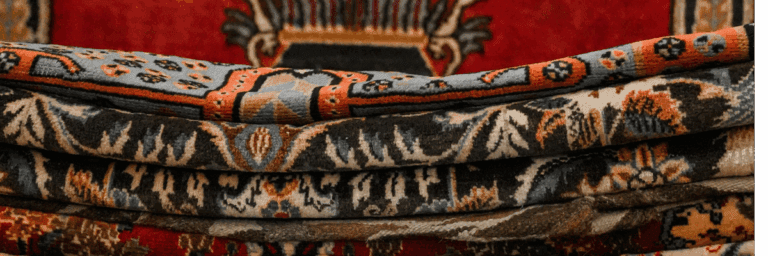 Stack of hand-knotted Persian carpets showing detailed floral patterns in coral, black, and cream colors