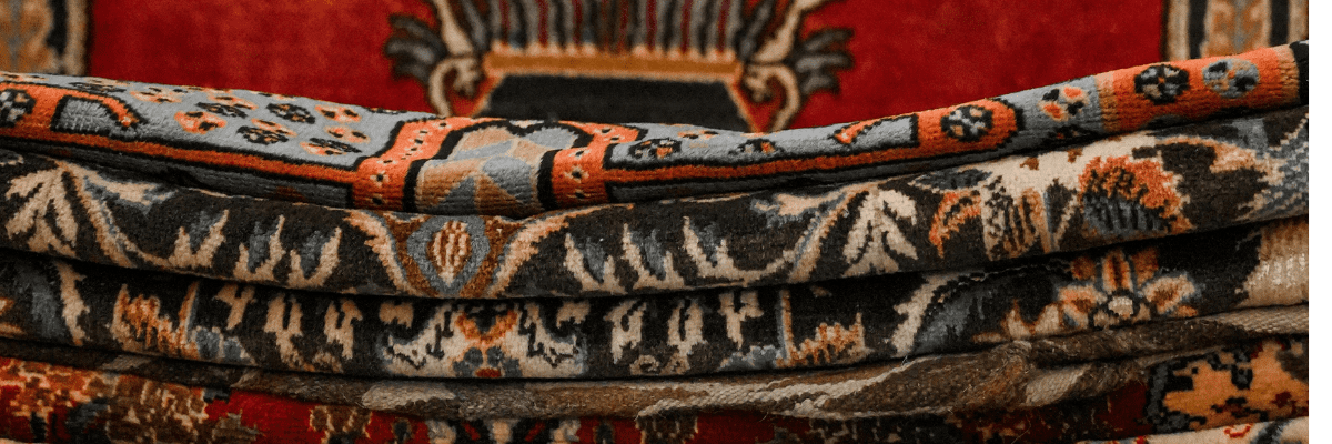 Stack of hand-knotted Persian carpets showing detailed floral patterns in coral, black, and cream colors